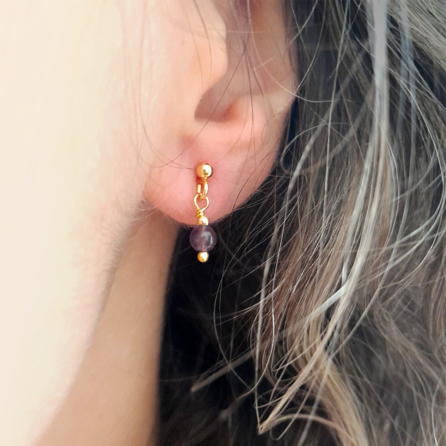Close-up of an ear adorned with tiny silver earrings featuring a single 4mm round amethyst gemstone dangling from a gold ball earstud.