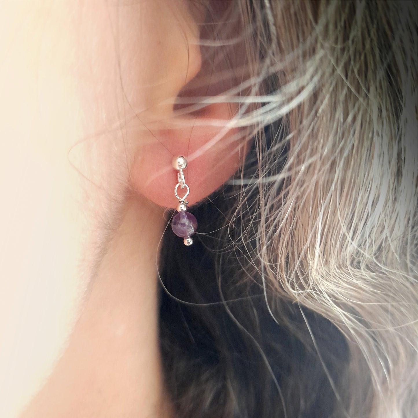 Close-up of an ear adorned with tiny silver earrings featuring a single 4mm round amethyst gemstone dangling from a silver ball earstud.