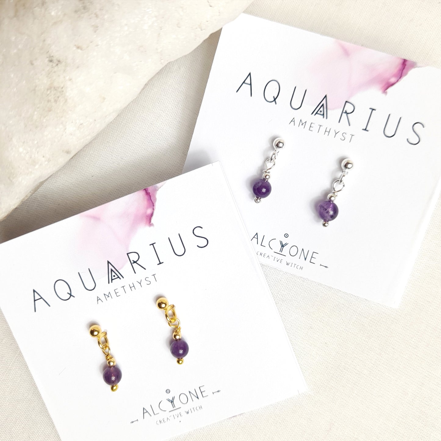 Two pairs of tiny and cute amethyst earrings, one with silver and the other with gold. Each features a single 4mm round amethyst gemstone dangling from a silver or gold ball earstud. Presented on two small square cards with 'Aquarius' and 'Amethyst' written on them, along with the Alcyone Jewellery logo