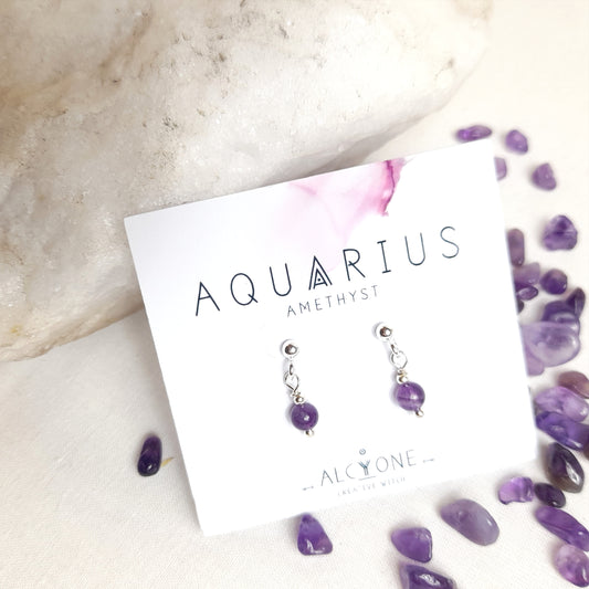 Tiny and cute silver earrings featuring a single 4mm round amethyst gemstone dangling from a silver ball earstud. Presented on a small square card with 'Aquarius' and 'Amethyst' written on it, along with the Alcyone Jewellery logo.